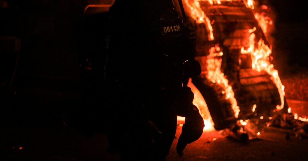 A tense night scene in Bogotá with a silhouette of a police officer against a fire backdrop during protests.